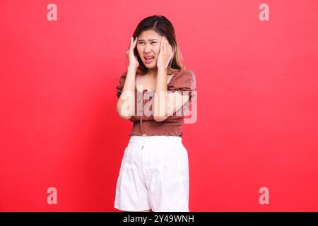 Expression de femme asiatique avec les deux mains tenant la tête, très douloureuse, vertigineuse, franche, portant une blouse brune et un short avec un fond rouge. pour h. Banque D'Images