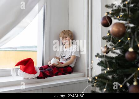 Enfant assis sur le rebord de la fenêtre près de la fenêtre et de l'arbre de Noël. Sur les pieds du garçon est chapeau de Père Noël rouge. Photos confortables festives sur un thème du nouvel an. concept de prépari Banque D'Images