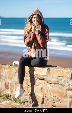 Une femme en veste à capuche assise au bord de la mer un jour ensoleillé Banque D'Images