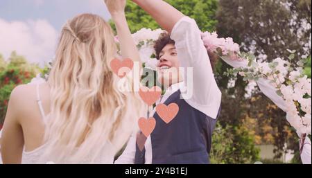 Image de coeurs roses sur un couple diversifié heureux dansant dans le jardin le jour du mariage Banque D'Images