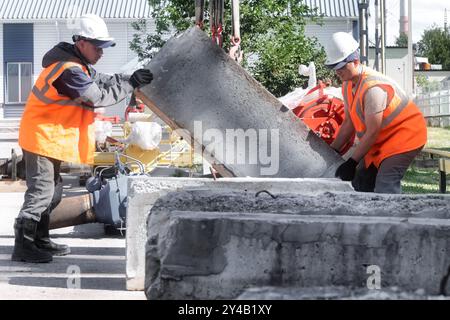 Deux ouvriers de la construction portant des équipements de sécurité soulevant et positionnant ensemble une dalle de béton lourde sur un chantier de construction occupé rempli de divers outils et matériaux. Banque D'Images