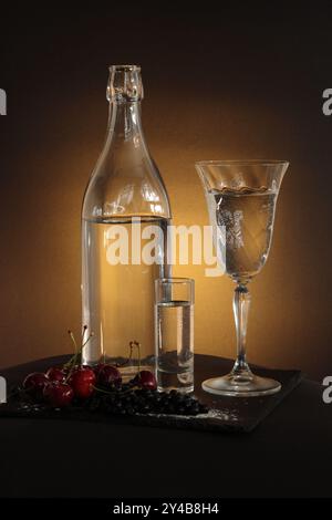 Cerises, verre et plan de liquide clair dans une nature morte magnifiquement éclairée. La lueur jaune contraste l'obscurité, apportant de la profondeur à la composition. Banque D'Images