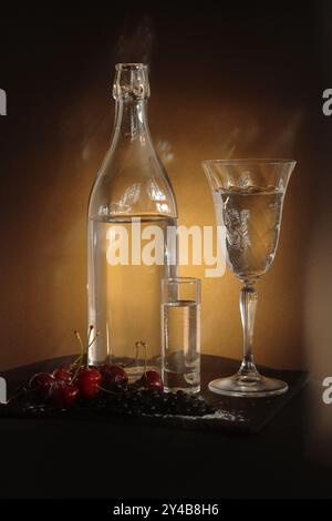 Cerises mûres à côté du verre, de la grenaille et du gobelet. Le triangle de lumière crée un effet dramatique sur la toile de fond sombre, soulignant la fraîcheur. Banque D'Images
