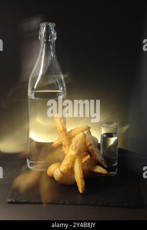 Un verre et une dose de liquide clair à côté de pommes de terre de style rustique sur un fond sombre. Le triangle de lumière ajoute de la chaleur et contraste le cadre sombre Banque D'Images