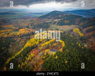 Un paysage d'automne à couper le souffle présente des teintes brillantes d'orange, de jaune et de rouge parmi les collines vallonnées. Une route sinueuse traverse la forêt colorée, invitant à l'exploration dans ce cadre serein. Banque D'Images