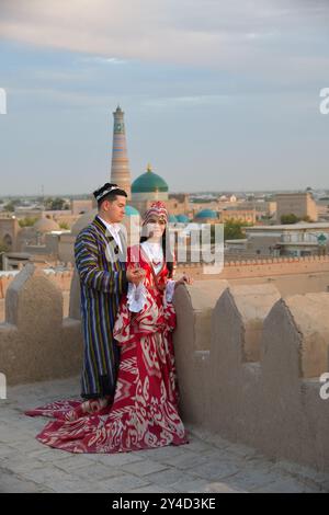 Khiva, Ouzbékistan - 15 septembre 2024 : jeune couple non identifié en tenue traditionnelle sur fond d'horizon ancienne ville de Khiva et Islam Khodja Mina Banque D'Images