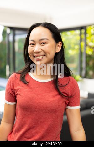 Jeune femme asiatique souriante dans une chemise rouge décontractée debout à l'intérieur, regardant la caméra Banque D'Images