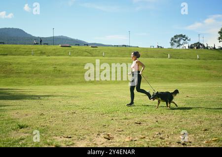 Jeune femme active faisant du jogging avec son chien à travers un champ herbeux ouvert par une journée ensoleillée. Concept de style de vie sain et de bien-être Banque D'Images