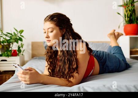 Une jeune et belle femme grande taille se prélasse sur son lit, absorbée dans son téléphone, entourée d'une atmosphère sereine de maison. Banque D'Images