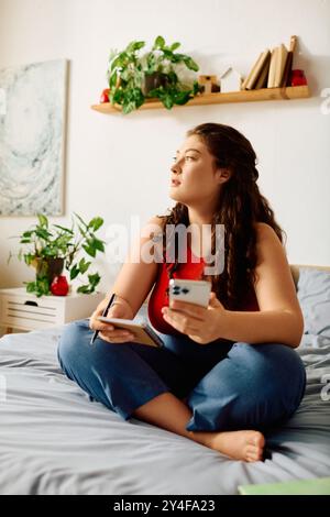 Une jeune femme de grande taille se détend sur son lit, notant des pensées tout en étant entourée d'un décor chaleureux et de plantes. Banque D'Images