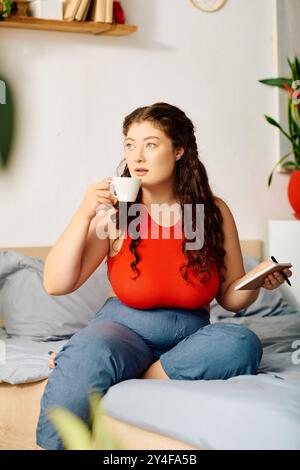 Une jeune femme de grande taille aux cheveux bouclés est assise sur son lit, sirotant du thé et notant des pensées dans son journal, embrassant un moment de tranquillité. Banque D'Images