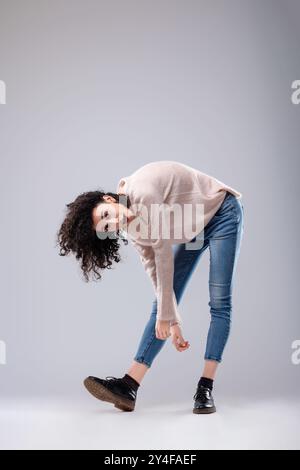 Jeune femme aux cheveux bruns bouclés pose de manière ludique dans un studio, exsudant confiance en soi et attitude insouciante Banque D'Images