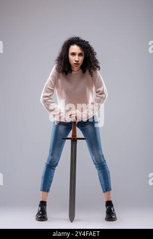 Modèle de mode confiant avec cheveux bouclés pose avec épée, exsudant détermination et force. Embrassant le pouvoir des filles, elle met en valeur le style tendance et Uni Banque D'Images