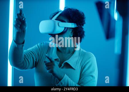 Portrait de vue latérale de femme noire portant un casque VR dans le spectacle d'art moderne interagissant avec des néons bleus expérimentant le cyberespace numérique Banque D'Images