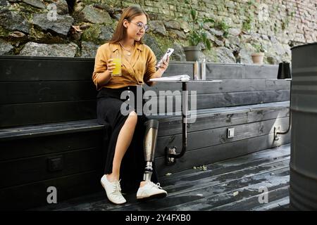 Une jeune femme avec une jambe prothétique se détend dans un café, sirotant un verre et vérifiant son téléphone, incarnant la vie quotidienne. Banque D'Images