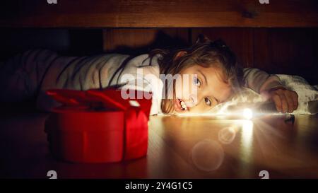 Petite fille allongée sur le sol dans la chambre et regardant sous le lit avec lampe de poche. Enfant excité et joyeux trouvant une boîte de cadeau rouge Banque D'Images