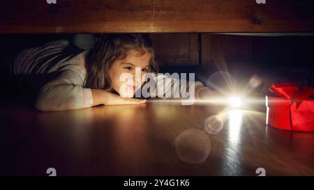 Petite fille allongée sur le sol dans la chambre et regardant sous le lit avec lampe de poche. Enfant excité et joyeux trouvant une boîte de cadeau rouge. Banque D'Images