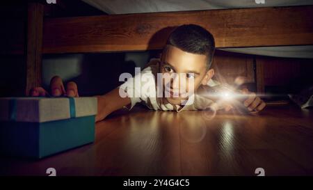 Petit garçon émotionnel, enfant allongé sur le sol dans la chambre et regardant sous le lit avec une lampe de poche. Enfant excité et joyeux trouvant la boîte de cadeau. Banque D'Images