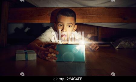 Petit garçon émotionnel, enfant allongé sur le sol dans la chambre et regardant sous le lit avec une lampe de poche. Enfant excité et joyeux trouvant la boîte de cadeau. Banque D'Images