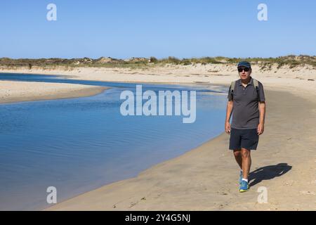 Un homme marche le long d'une plage de sable, avec un sac à dos à la main. Il est habillé de façon décontractée en short, un polo et une casquette, suggérant une promenade tranquille Banque D'Images