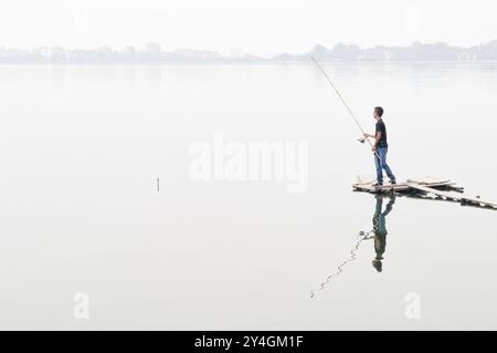 Pêcheur pêchant sur le lac de l'Ouest Hanoi Vietnam // HANOI, Vietnam — Un pêcheur jette sa ligne depuis une jetée en bois sur le lac de l'Ouest (Ho Tay), le plus grand lac d'eau douce de Hanoi. Une brume épaisse obscurcit le rivage lointain du lac de 500 hectares. Les préoccupations environnementales persistent au sujet de la qualité de l'eau et de la consommation de poisson des lacs urbains de Hanoi en raison des niveaux de pollution importants. Banque D'Images