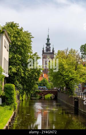 Vue panoramique sur le canal avec une végétation luxuriante et l'imposante tour de l'horloge de l'église de Catherine à Gdansk, Pologne, lors d'une journée paisible. Banque D'Images