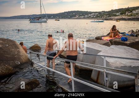 Deux Italiens d'âge moyen entrent dans la mer entre les rochers et à côté des défenses maritimes protégeant Castro Marina dans les Pouilles, en Italie. Banque D'Images