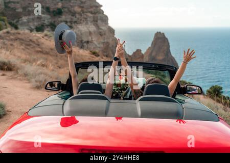 Une voiture cabriolet rouge avec deux personnes dedans, dont l'une ondule. La voiture roule sur une route près de l'océan. Banque D'Images