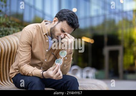 Jeune homme assis sur un banc à l'extérieur semblant frustré ou stressé. Il semble éprouver des maux de tête ou de l'inconfort dans un cadre détendu. Les émotions comprennent la frustration, la fatigue, le stress. Banque D'Images