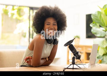 Femme afro-américaine souriante avec écouteurs et micro enregistrant podcast au bureau à domicile Banque D'Images