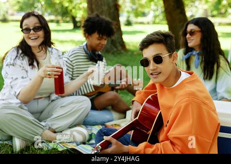 Plan moyen d'un adolescent avec des lunettes de soleil jouant de la guitare et chantant accompagné par des amis assis à proximité sur l'herbe ayant pique-nique dans le parc, espace de copie Banque D'Images