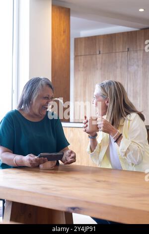Riant ensemble, amies féminines multiraciales âgées appréciant le café et la conversation à la maison Banque D'Images