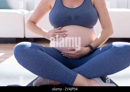 Femme enceinte assise sur un tapis de yoga, tenant le ventre et se relaxant à la maison Banque D'Images