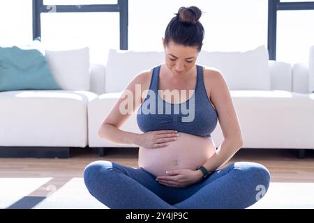 À la maison, femme enceinte assise les jambes croisées, tenant le ventre et méditant dans le salon Banque D'Images