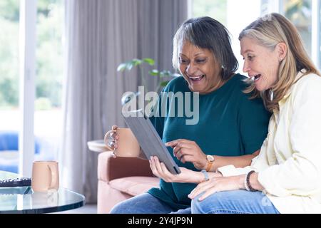 À la maison, en utilisant une tablette et en buvant du café, des amies féminines multiraciales âgées profitant du temps ensemble Banque D'Images