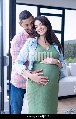 S'attendre à un couple multiracial embrassant, souriant et touchant le ventre enceinte dans une maison confortable Banque D'Images
