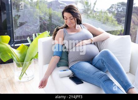 À la maison, femme enceinte mesurant la pression artérielle avec un moniteur numérique tout en se relaxant sur le canapé Banque D'Images