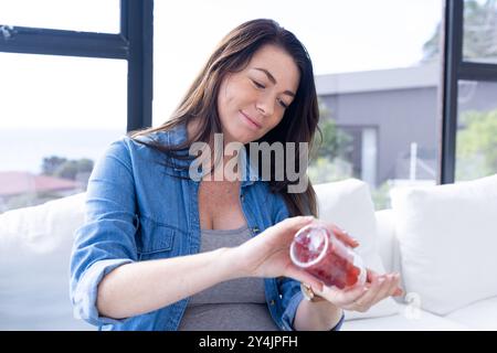 À la maison, femme enceinte tenant une bouteille de vitamines, souriant et assise sur le canapé Banque D'Images