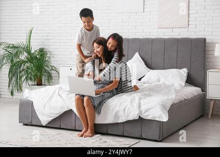 Belle jeune heureuse mère asiatique avec ses petits enfants mignons regardant des dessins animés sur ordinateur portable dans la chambre Banque D'Images