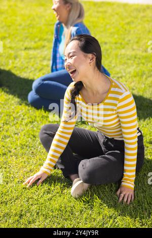 Assise sur l'herbe, jeune femme riant avec divers amis à l'extérieur, profitant d'une journée ensoleillée Banque D'Images
