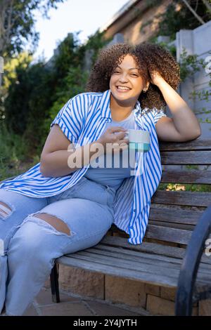 Femme souriante avec les cheveux bouclés assis sur le banc tenant la tasse de café profitant de la détente en plein air Banque D'Images