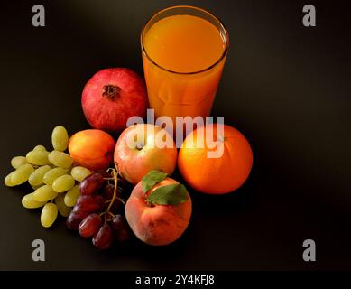 Un mélange de jus de fruits dans un grand verre à facettes sur un fond noir, à côté de fruits mûrs, pomme, orange, pêche, grenade et grappes de raisins. Banque D'Images