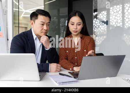 Professionnels asiatiques travaillant ensemble sur le projet dans un cadre de bureau moderne. Deux collègues se sont concentrés sur l'écran d'ordinateur portable, discutant de stratégie et prenant des décisions commerciales importantes. Banque D'Images