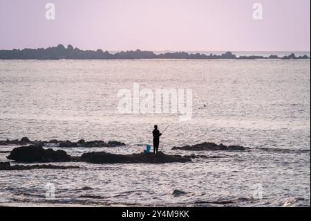Un pêcheur solitaire debout sur un éperon rocheux contre un coucher de soleil magnifique. La silhouette du pêcheur crée un sentiment de tranquillité et de solitude. S Banque D'Images