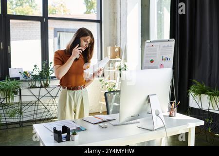 Une jeune personne non binaire se tient dans un bureau lumineux, multitâche avec des documents et un téléphone. Banque D'Images