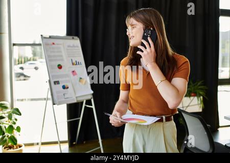 Une jeune personne non binaire est sur un appel téléphonique tout en analysant des graphiques et des données de bureau. Banque D'Images