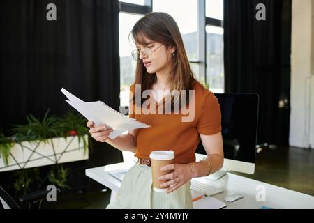 Une jeune personne non binaire se tient dans un bureau lumineux, lisant des notes avec une tasse à café à la main. Banque D'Images