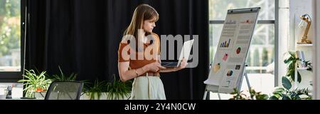 Un jeune individu non binaire analyse les graphiques tout en tenant un ordinateur portable dans un bureau élégant. Banque D'Images