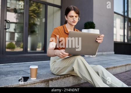 Engagé dans un travail ciblé, une jeune personne non binaire est assise à l'extérieur d'un bureau avec un ordinateur portable. Banque D'Images