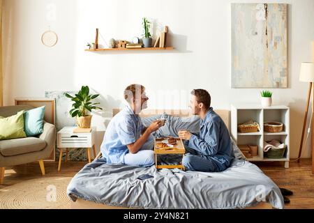 Un joyeux couple gay profite d'une matinée tranquille ensemble en pyjama, partageant le petit déjeuner et riant au lit. Banque D'Images